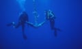 Two divers raise an anchor covered in rust and barnacles towards the surface