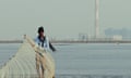 An Indian man with a fishing net by the sea, with a power plant in the distance behind him