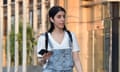 An Arab woman with her head uncovered walking down a street in a T-shirt and dungarees while listening to earphones