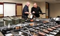 two men examine a gun inside a room with a table covered with guns