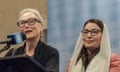 An older woman wearing black rimmed glasses and a black top speaks into a microphone while standing next to a smiling middle-aged woman in a white headscarf also wearing black rimmed-glasses.