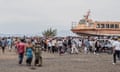 A crowd of people stand on the shore with an orange boat seen raised out the water.