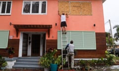 A man standing on a ladder fixes boards over windows of a house as another holds the foot