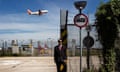 A black man in a black suit, tie and sunglasses, and with a serious demeanour, stands at a gate on the outskirts of an airport