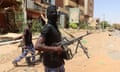 A man in a balaclava holding a rifle looks back at the camera while walking along a dirt street