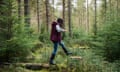 A mid adult woman wearing wellington boots, walking in Thrunton Woods, Northumberland. She is stepping onto a tree stump from a fallen log with her arms outstretched to balance herself.