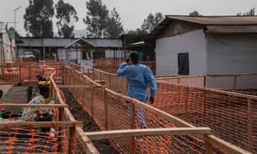 A man wearing blue PPE overalls walks between barriers made of orange netting and wood past roughly constructed buildings made of wood with tin roofs.